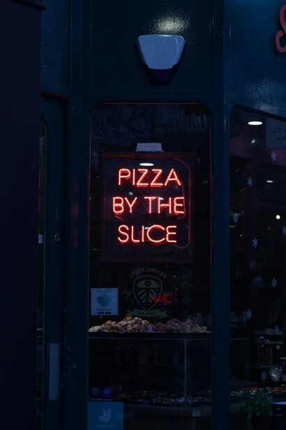 An image of a neon sign at night that says, "Pizza By The Slice."